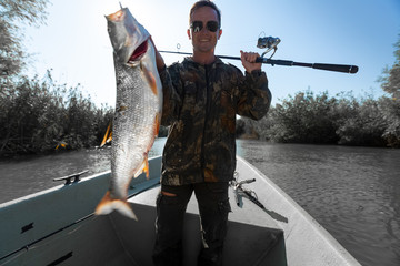 Happy fisherman holds trophy Asp fish being in the boat on a lake. Fish is motion blurred