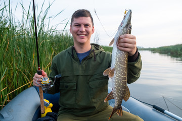 Happy fisherman holds pike fish being in the boat on the lake