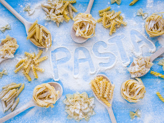 Fototapeta na wymiar Word Pasta on wooden table with variety of types and shapes of dry Italian pasta and flour. Pasta ingredients on wooden spoons and blue background. Top view Flat lay Colorful and stylish composition.