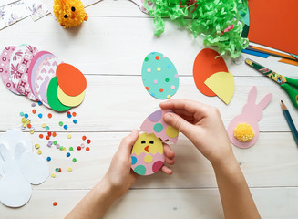 The child makes crafts with his own hands for the Easter. Colorful handmade from multi-colored paper. Scissors, cardboard, eggs, chicken, rabbit. Art creativity on a wooden table. Top view, copy space