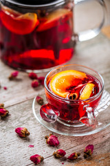 Appetizing healthy herbal red berry tea with orange in a glass teapot and cup on the wooden background decorated with roses, vertical