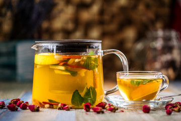 Closeup on the healthy herbal yellow apple tea with lemon and cinnamon in a glass teapot and mug on the wooden background decorated with roses, horizontal