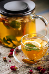 Closeup on the healthy herbal yellow apple tea with lemon and cinnamon in a glass teapot and mug on the wooden background decorated with roses, vertical