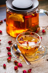 Closeup on the healthy herbal yellow apple tea with lemon and cinnamon in a glass teapot and mug on the wooden background decorated with roses, horizontal