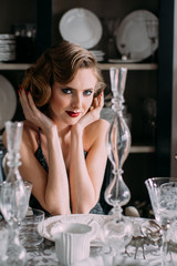 Beautiful blond girl with vintage make up and hairstyle in evening shining dress sitting at table with plates and glasses