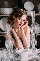 Beautiful blond girl with vintage make up and hairstyle in evening shining dress sitting at table with plates and glasses
