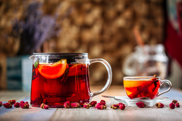 Appetizing healthy herbal red berry tea with orange in a glass teapot and cup on the wooden background decorated with roses, horizontal