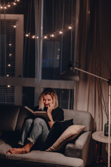 Girl reads book in the evening on couch. Night lamp light. Relaxing evening. Large window. Modern...