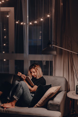 Girl reads book in the evening on couch. Night lamp light. Relaxing evening. Large window. Modern...