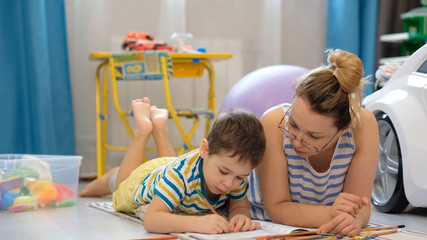 Young happy mother and little son lies on the floor in the children's room and doing arts and crafts, drawing together