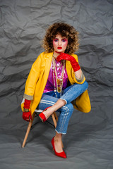 A girl in a yellow jacket and blue jeans with an afro hairstyle sits on a chair. Fashion eighties, the era of disco. Studio photo on a gray background.
