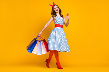 Full size photo cheerful girl shopping center client buy bargain hold bags go step use smartphone wear dotted blue skirt tights red headband high-heels isolated shine yellow color background