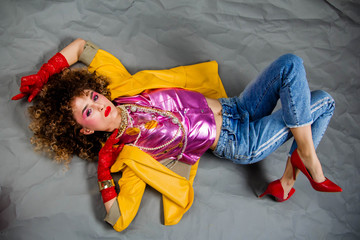 The girl in a yellow jacket and blue jeans with an afro hairdo lies on the floor. Fashion eighties,...