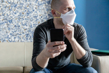 young man with ffp1 respirator on self-isolation in his apartment during quarantine of coronavirus covid-19 reads news on a smartphone