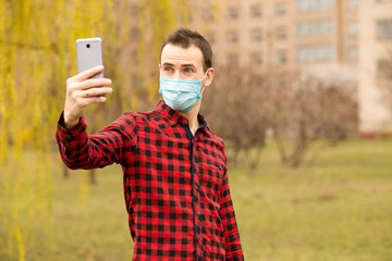 Happy young man in sterile face mask making self photo or video call with mobile phone show hello gesture walking by street.