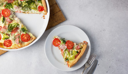 A portion of open pie with broccoli, tomato and bacon on a white plate and quiche in a baking dish nearby.