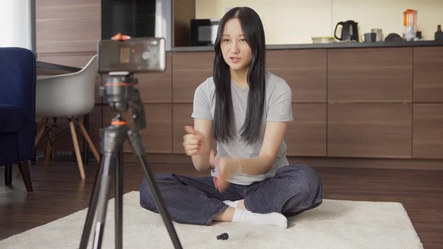 Female Asian lifestyle blogger sitting cross-legged on floor at home and spraying antiseptic sanitizer on hands while recording video on cell phone placed on tripod