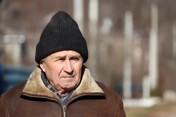 Elderly man standing on rural background in sunny spring day. Sad face expression, concept of walking during coronavirus quarantine, life in village, old age