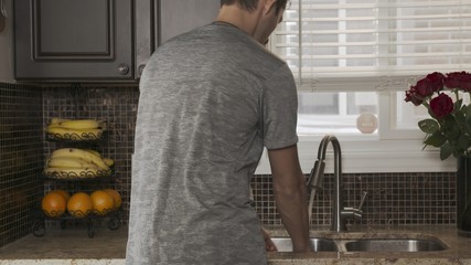 young attractive man in a gray t-shirt stands with his back to the camera and washes dishes or hands in a beautiful fashionable clean kitchen in the sink. A guy washes vegetables and fruits in the