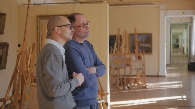 Two men in an art gallery look at a painting and talk about art