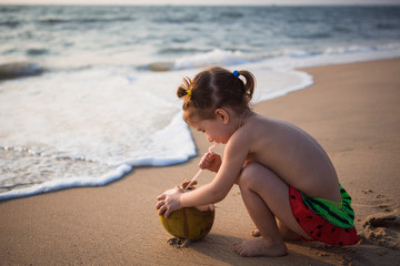 A little girl in a watermelon swimsuit squats on the beach near the sea and plays with a drinking coconut. A girl plays on the sand with a coconut and a straw.