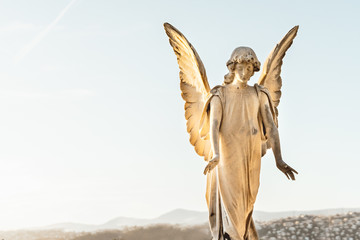 statue of angel with wings against light sky at cemetery. Closing stoned angel in an old cemetery.. Graveyard old weathered stone sad angel sculpture on funeral. copy space text.