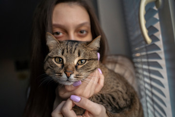 A girl in a black medical mask by the window hugs a brown cat. Pets. Self isolation.