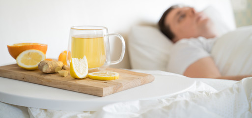 Obraz na płótnie Canvas Cup with antipyretic drugs for colds,flu.Sick man in bed. Tea with citrus vitamin C,ginger root,lemon,orange.Wooden tray. Home self-treatment.Medical quarantine antiviral covid-19 coronavirus therapy