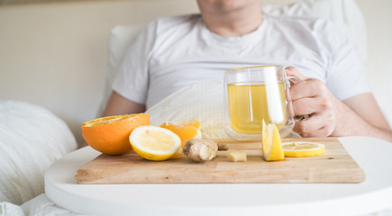 Cup with antipyretic drugs for colds,flu.Sick man in bed. Tea with citrus vitamin C,ginger root,lemon,orange.Wooden tray. Home self-treatment.Medical quarantine antiviral covid-19 coronavirus therapy