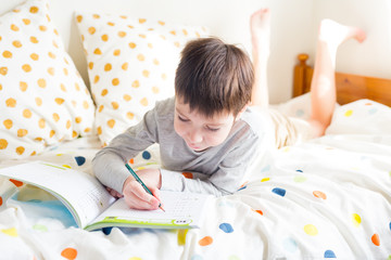  Distance learning online education. School Boy on bed and doing school homework at home, writing in notebook. Quarantine. Concept staying at home during emergency