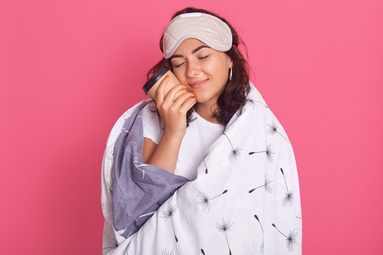 Close up portrait of dark haired woman posing with closed eyes, holding takeaway hot coffee, enjoying stay home during quarantine, wrapped in white blanket, isolated over pink stdio background.