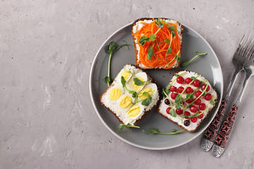 Three sandwiches on toast with cream cheese, carrots, red currant and quail eggs decorated with microgreens peas on a plate on gray concrete background, Copy space, View from above