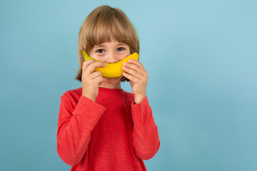 sweet european boy in red sweater with banana in hands posing on isolated blue studio background