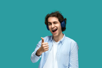Portrait of a young beautiful man wearing white t-shirt and blue shirt in headphones shows thumb up for approval and wink looking at camera over blue background