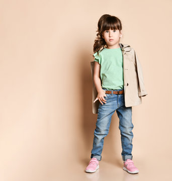 A Little Brunette Girl In A Beige Raincoat Holds Her Hand In The Pocket Of Her Jeans.