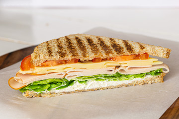 Club sandwich with ham, cheese, lettuce, and tomato served on white paper on wooden cutting board. Tasty homemade lunch, breakfast or take away food. 