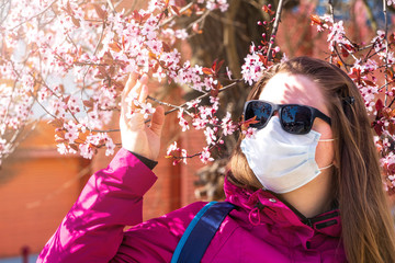 Girl or young woman in a medical mask and sunglasses sniffs flowers on blooming cherry or Apple...
