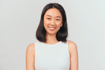 Portrait of girl tilts head and smiles sweetly showing white teeth. Beautiful young woman Asian appearance with black hair brown eyes stands isolated white background in Studio