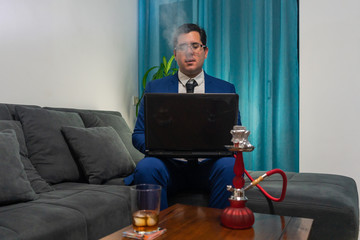 Young man sitting on a sofa. He is wearing a blue suit. He is working with his laptop. On a wooden table there is a glass of whiskey and a red shisha.