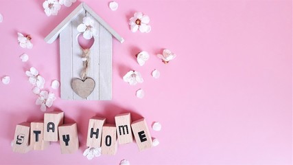 Wooden decorative house and wooden letters with text stay home,stey safe among apricot flowers on a pink background.Copy spaes.