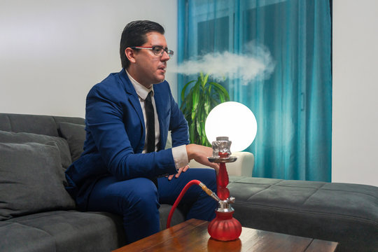 Young man sitting on a sofa. He smoking with a shisha. He is wearing a blue suit. The hookah is red.
