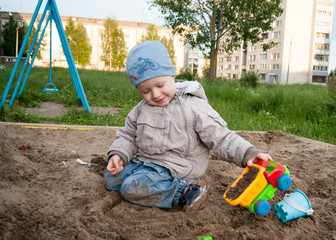 A fair-haired boy of three years in a blue hat and jeans, in a light jacket plays in the sandbox with toys