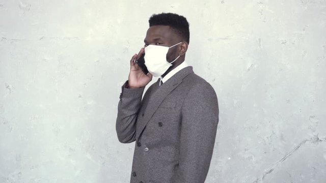 Side view of african man in a suit and medical mask talking by the phone. Black man stops against the concrete wall, waves his hand, and walks away, continuing the conversation. Coronavirus, epidemic