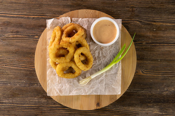 fried onion rings with sauce and green leek on paper and circle wooden board top view