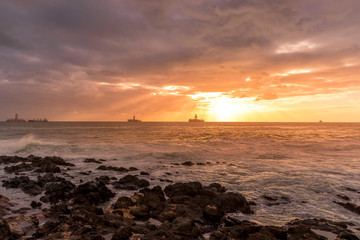 Fototapeta premium Sunrise from a stony shore to boats moored on the horizon with the rising sun and pesky shining through the clouds to the ocean with moving clouds from the Grand Canary Island.