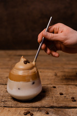Dalgona coffee cold drink with milk whipped with foam, instant coffee, Korean trend home-made. On the texture background of wood with a hand and eco friendly straw, vertically with space for text.