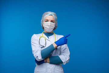Portrait of caucasian female with short dark hair in medical gown, blue medical gloves and special mask, a phonendoscope around her neck recommends to stay home