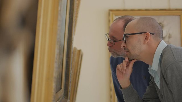 Two men in an art gallery look at a painting and talk about art