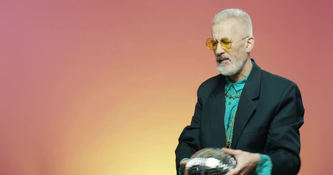 Stylish funny old Caucasian retired man in glasses dancing and having fun at 70s style party while holding silver disco ball on pink wall background. Male pensioner dancer.