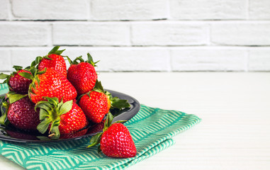 Red strawberries in a black plate with green kitchen towel close up on a white brick background with copy space